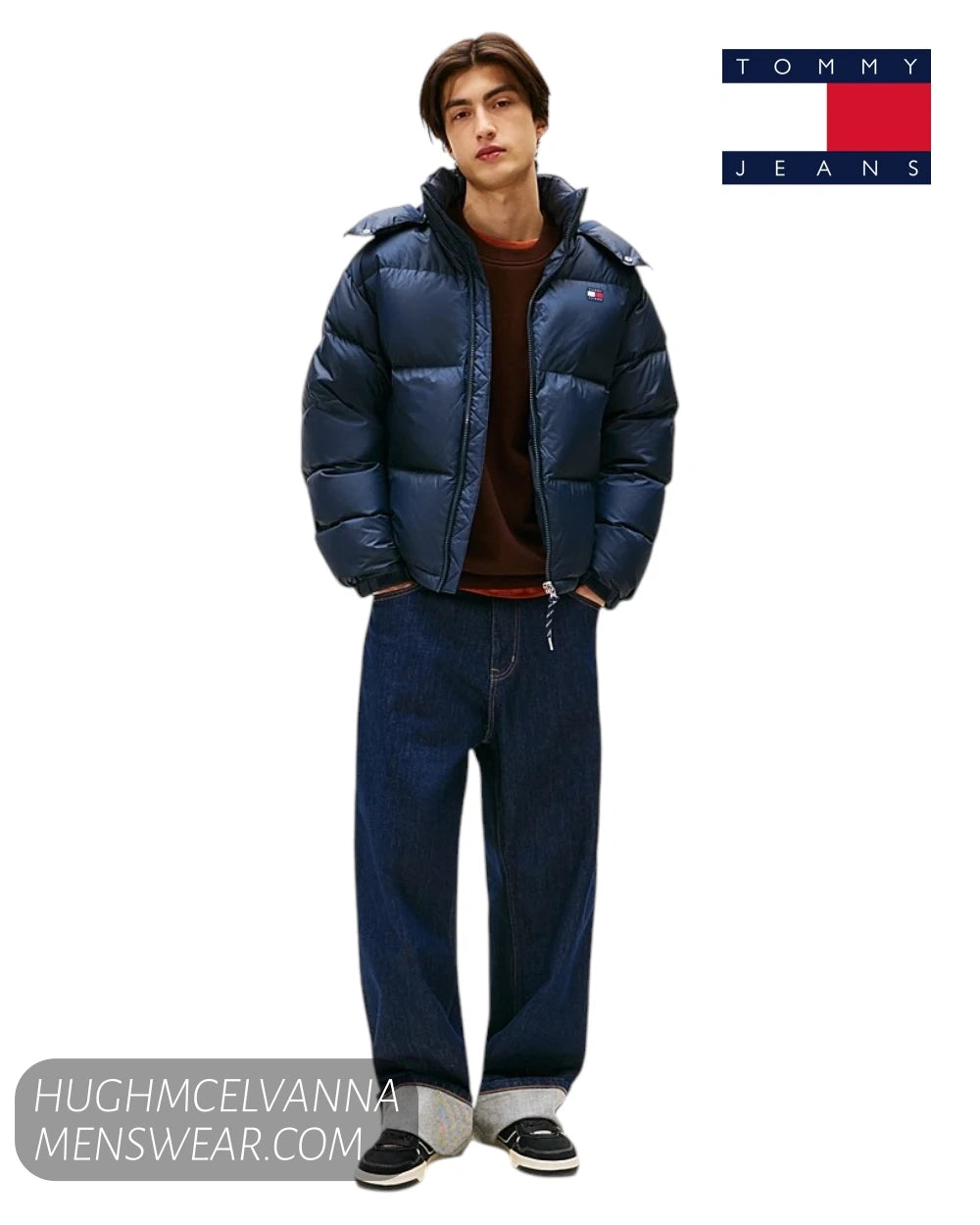 A model wearing a navy blue puffer jacket by Tommy Jeans, paired with dark brown and blue jeans, standing against a white background with the Tommy Jeans logo in the top right corner.