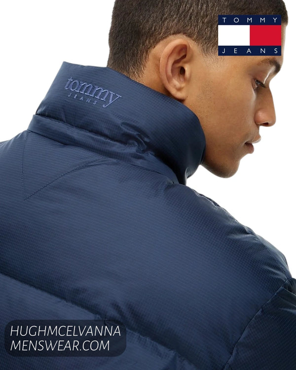 A close-up side profile of a man wearing a navy blue puffer jacket with a high collar, featuring the Tommy Jeans logo.