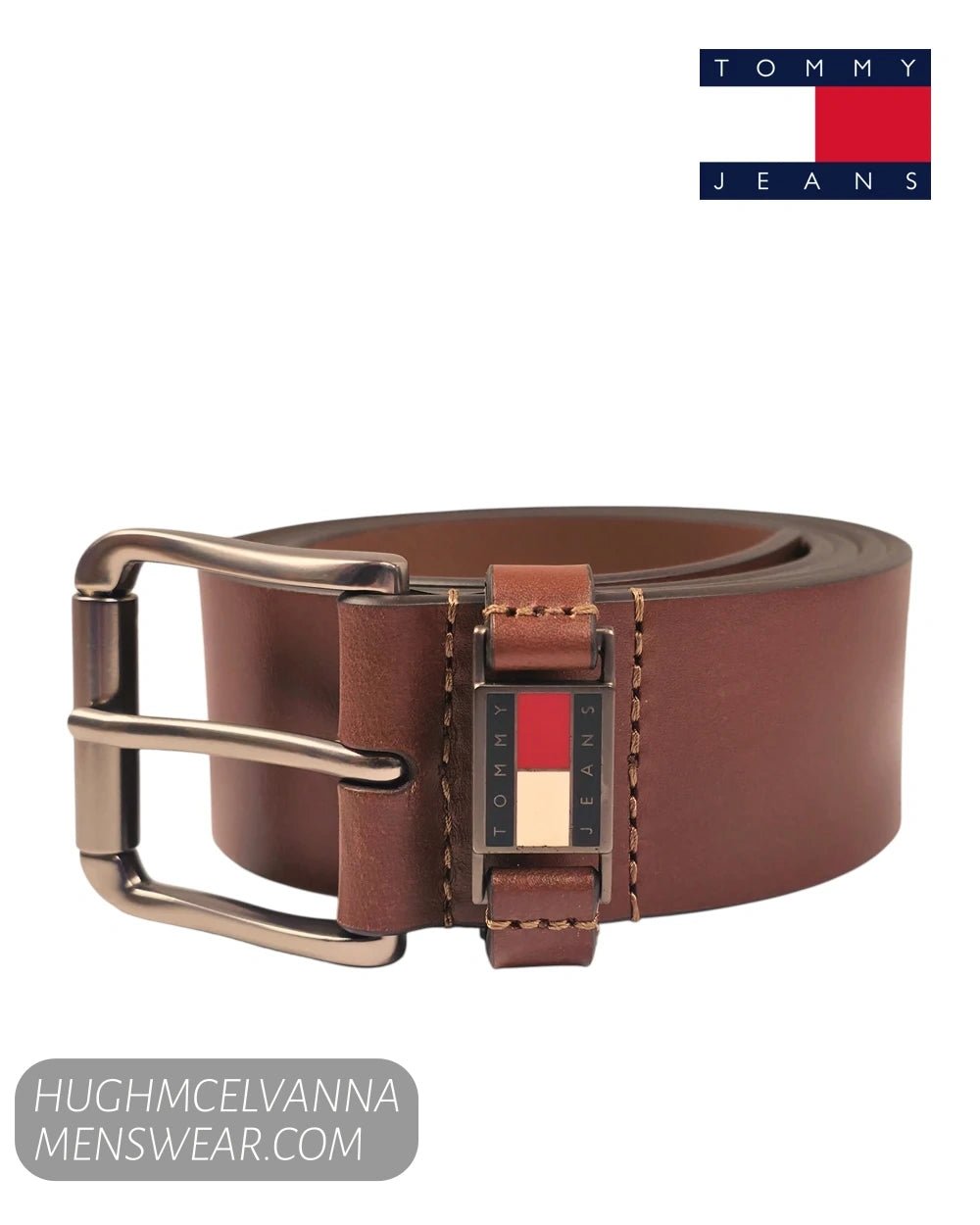 A brown leather belt with a silver-tone buckle and a signature Tommy Hilfiger flag emblem on the belt keeper, shown on a white background with a Tommy Jeans logo in the top right and a merchant watermark in the bottom left.