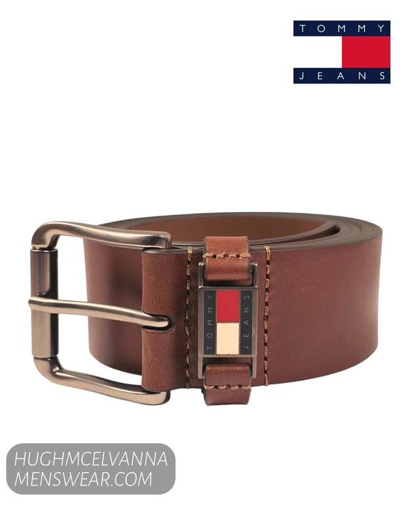 A brown leather belt with a silver-tone buckle and a signature Tommy Hilfiger flag emblem on the belt keeper, shown on a white background with a Tommy Jeans logo in the top right and a merchant watermark in the bottom left.