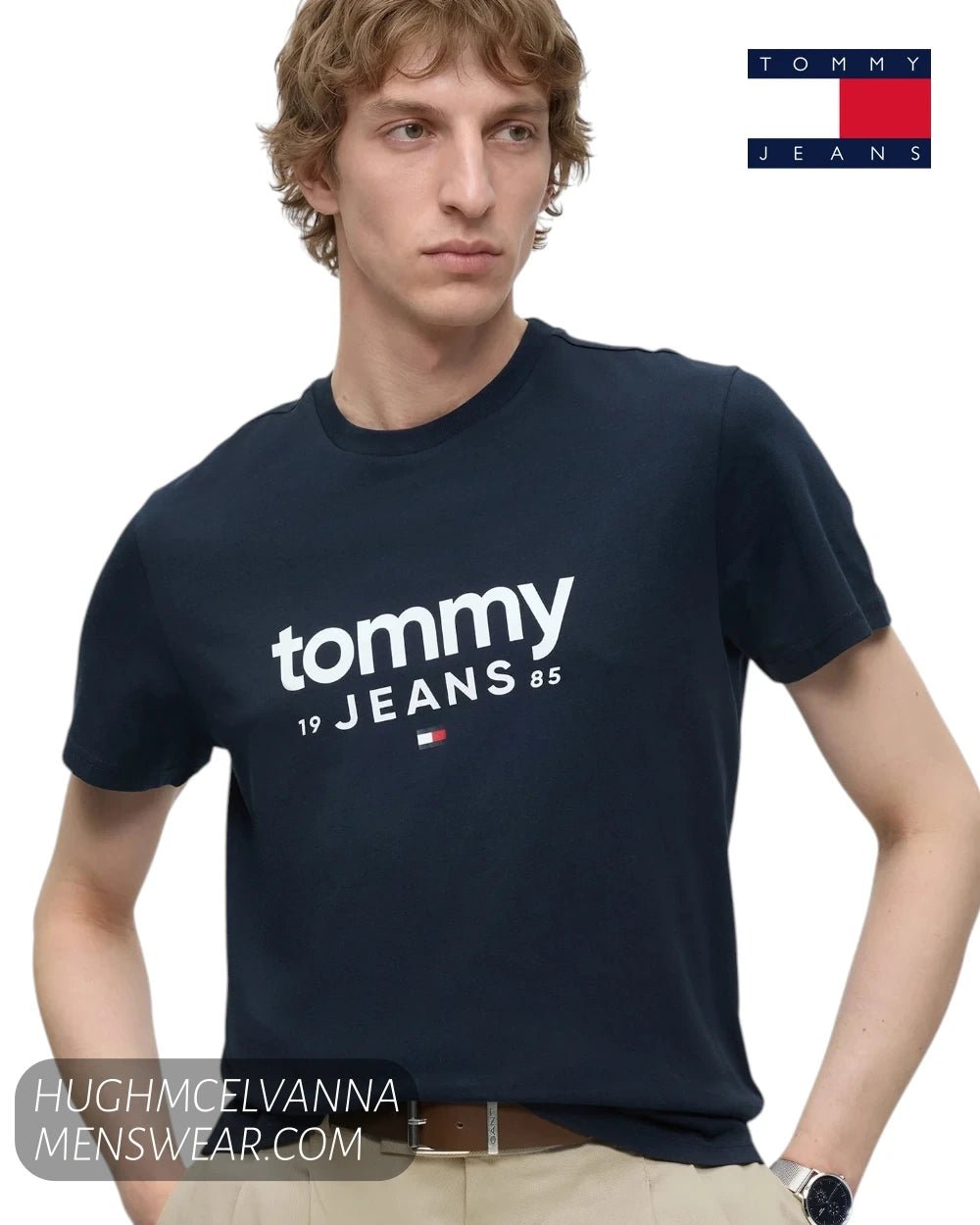 Front view of model wearing Tommy Jeans navy graphic logo t-shirt tucked into pleated beige trousers with leather belt