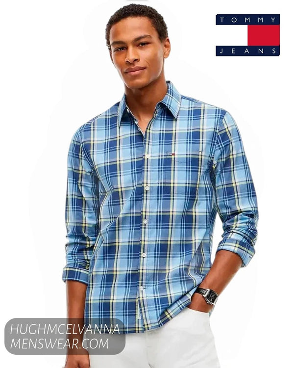 A man wearing a blue and white plaid long-sleeve button-down shirt by Tommy Jeans, paired with white trousers. 